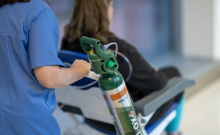 A hospital worker pusing a wheel-chair with a Walk-O2-Bout+ Medical gas cylinder attached