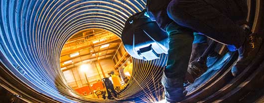 Welder standing inside of a large corrugated metal pipe performing a weld on the interior of the pipe underneath.